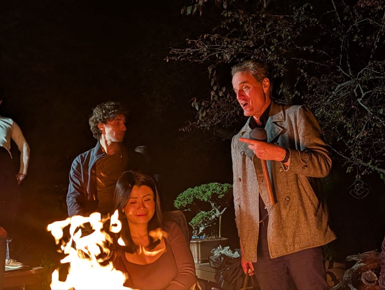 Speaker addressing the intimate circle around dancing flames with bonsai tree in background